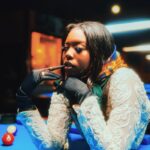 Elegant woman in fashion attire poses at a pool table in a dimly-lit bar setting.