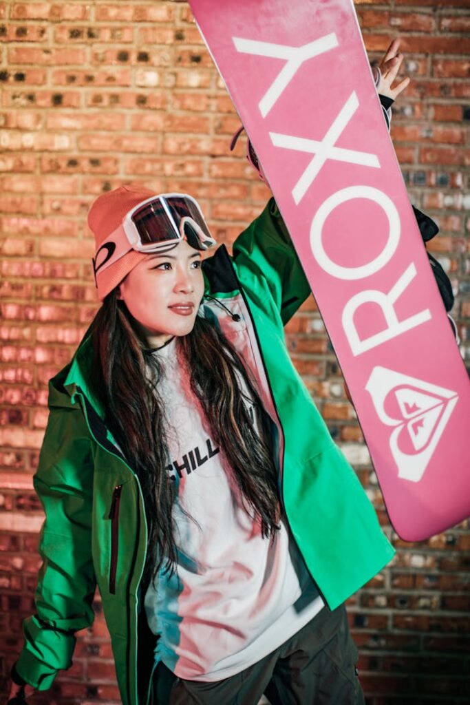 Asian woman in vibrant winter sportswear holding a snowboard against a brick wall.