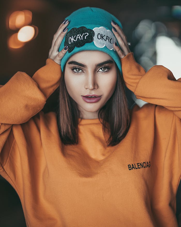 Stylish woman wearing an orange sweater and blue beanie posing confidently indoors.