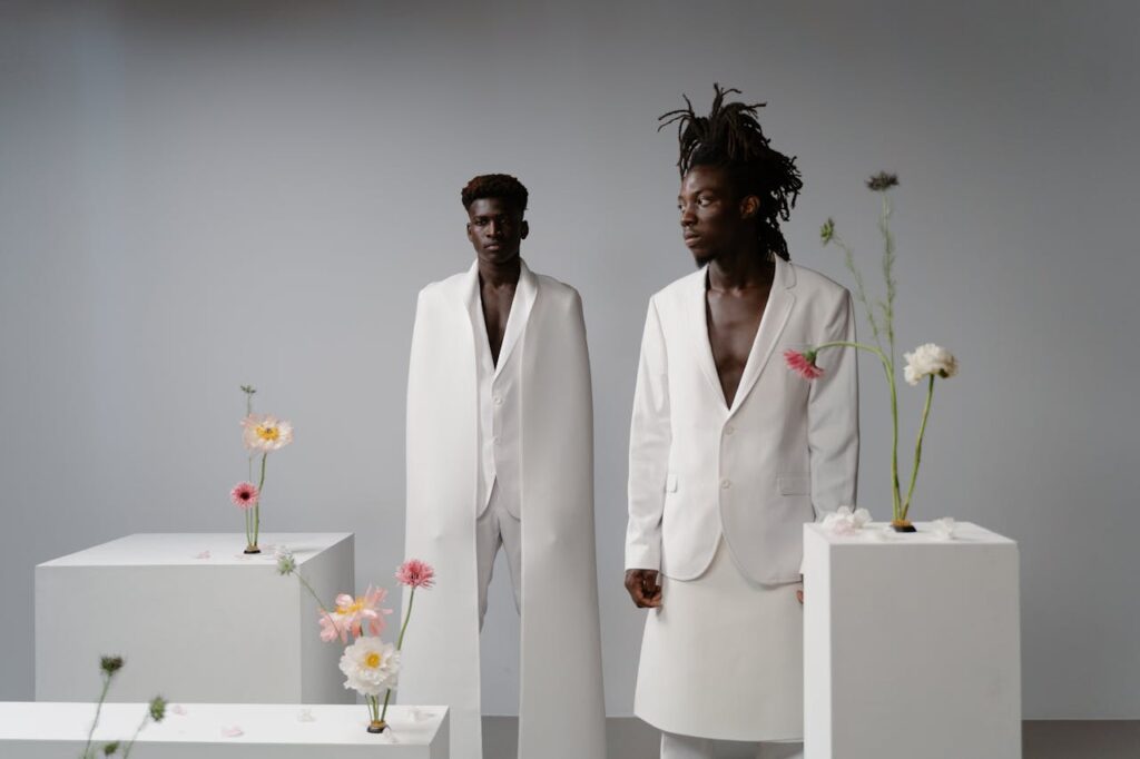 Stylish men in white suits pose amid floral arrangements in a minimalist studio setting.